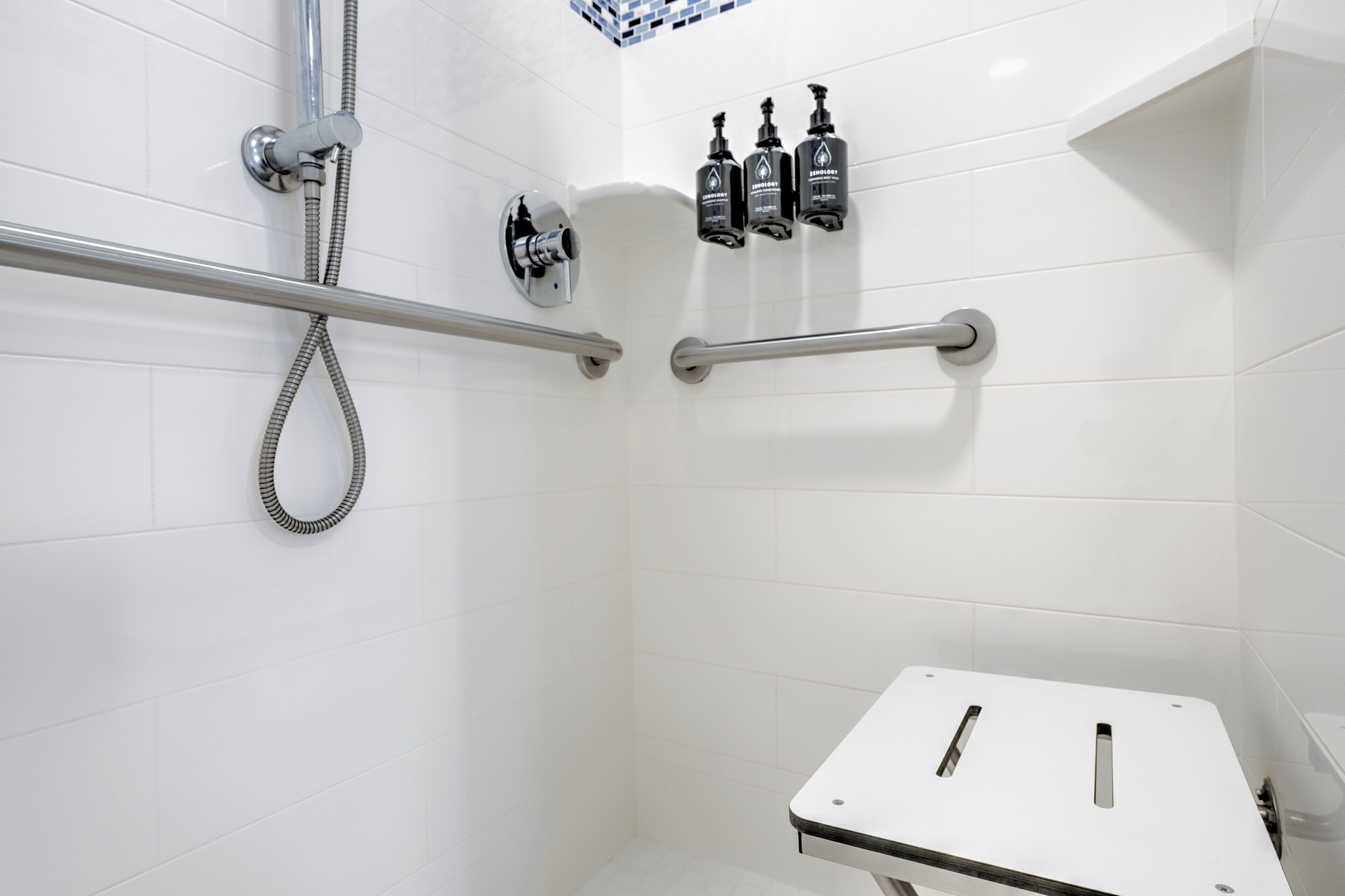 A modern bathroom with white tiles, a shower with a handheld showerhead, and three bottles of shampoo and conditioner on the wall.