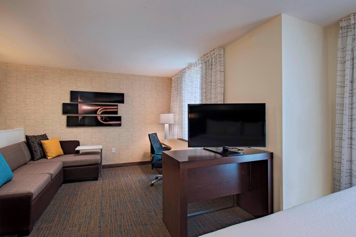 A hotel room with a sofa, a desk, a flat-screen TV, a chair, and a patterned carpet.