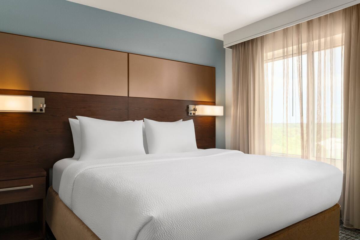 Habitación de hotel con una cama grande, cabecero de madera, dos lámparas de noche y una ventana con cortinas traslúcidas.