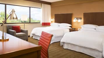 A hotel room with two beds, a desk, a chair, a lamp, and a view of trees through the window.