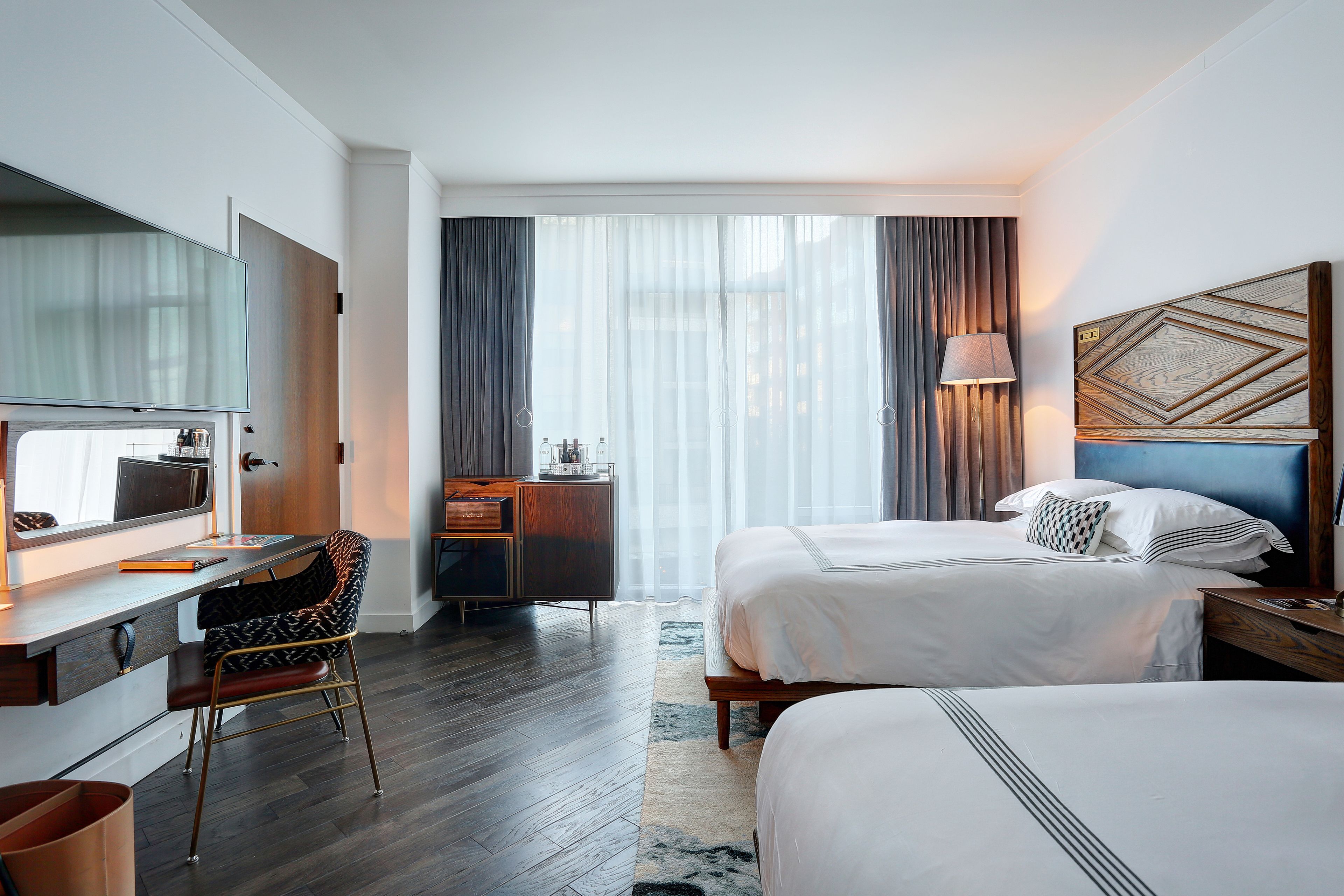 Ein Hotelzimmer mit zwei Betten, einem Schreibtisch, einem Sessel, einem Fernseher und einem großen Fenster mit Vorhängen.