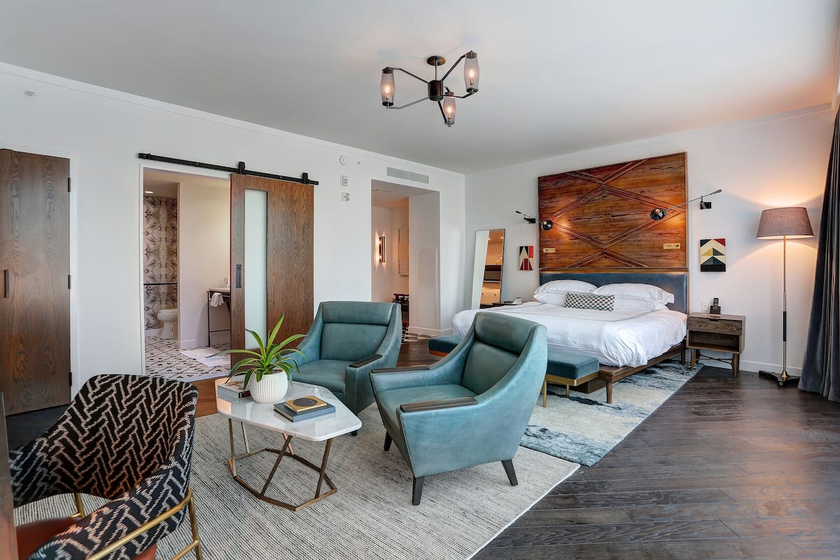Une chambre d’hôtel moderne avec un grand lit, deux fauteuils, une petite table avec une plante, une grande peinture abstraite au mur et un lampadaire.