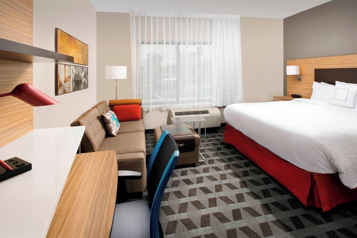 Ein Hotelzimmer mit einem Bett, einem Schreibtisch, einem Stuhl, einer Couch und einem Fenster mit durchsichtigen Vorhängen.