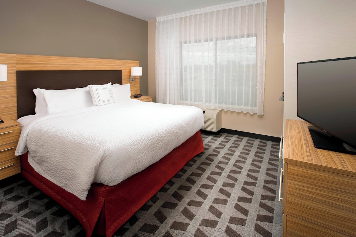 Ein Hotelzimmer mit einem großen Bett, einem Fernseher und einem Fenster mit durchsichtigen Vorhängen.