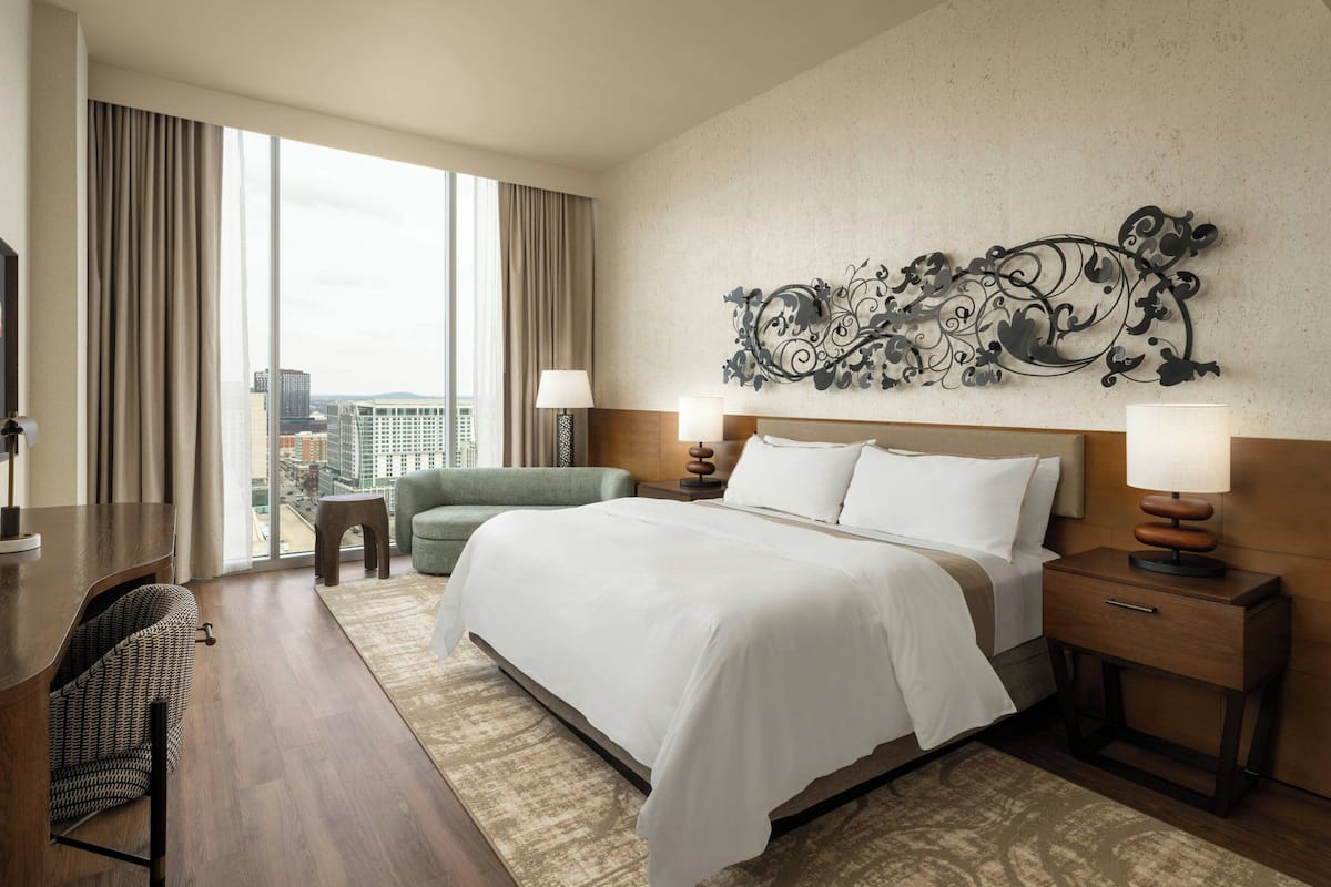 A hotel room with a large bed, two bedside tables with lamps, a desk with a chair, a sofa, and a large window with curtains.