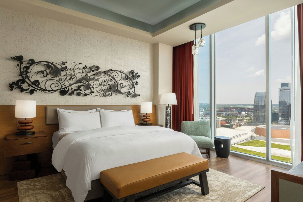 A hotel room with a large bed, two bedside tables, a bench, a chair, and a view of a cityscape through a large window.