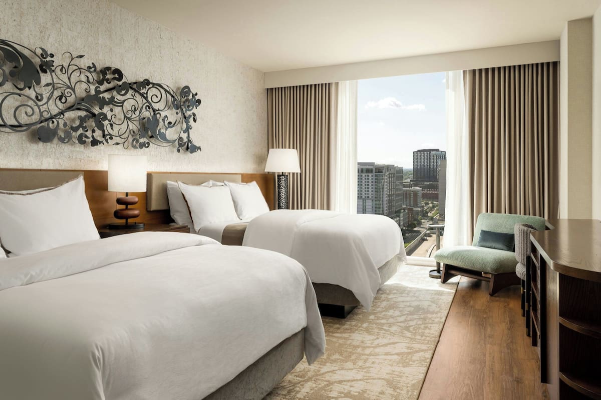 A hotel room with two beds, a desk, a chair, and a large window with a city view.