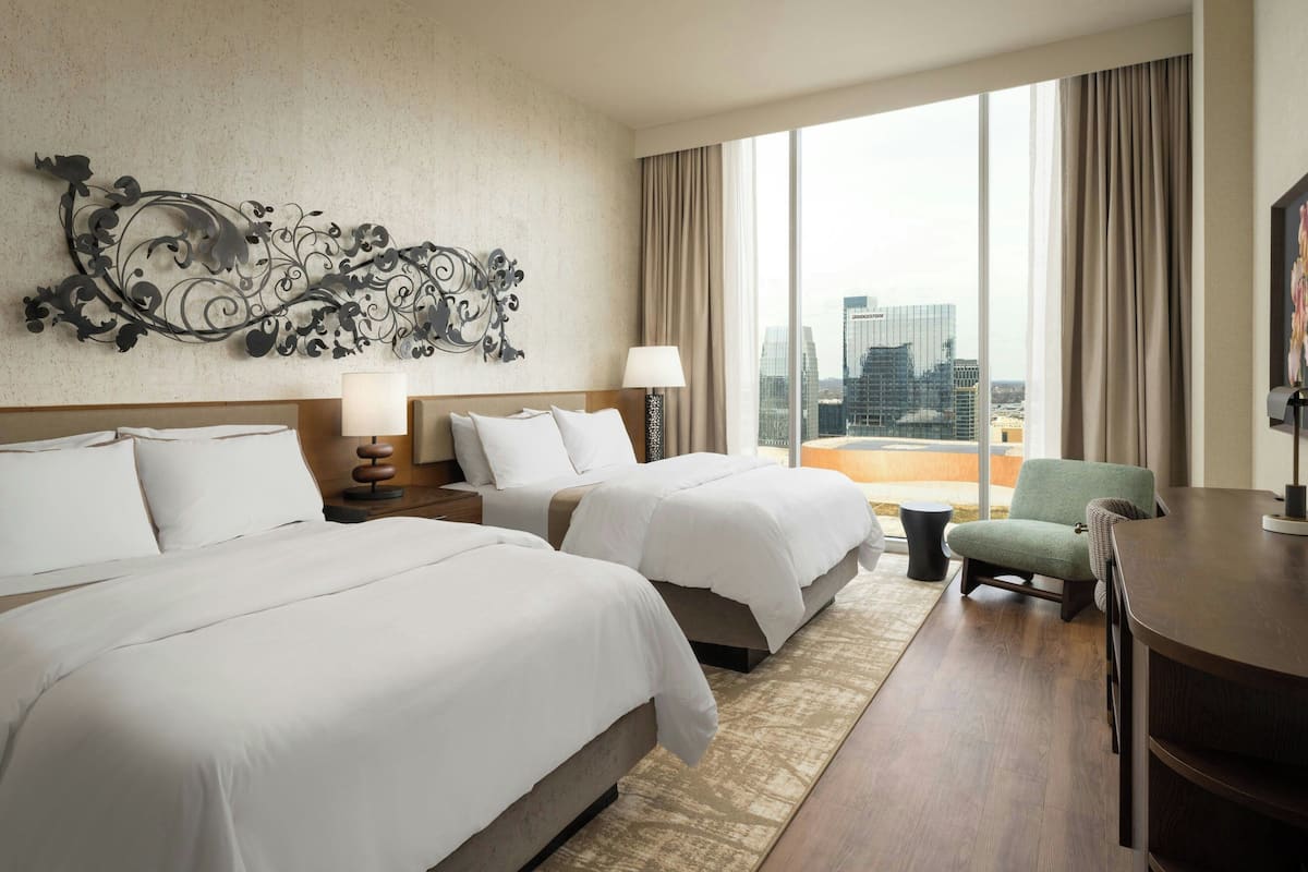 A hotel room with two beds, a desk, a chair, a large window with a city view, and a decorative wall piece.