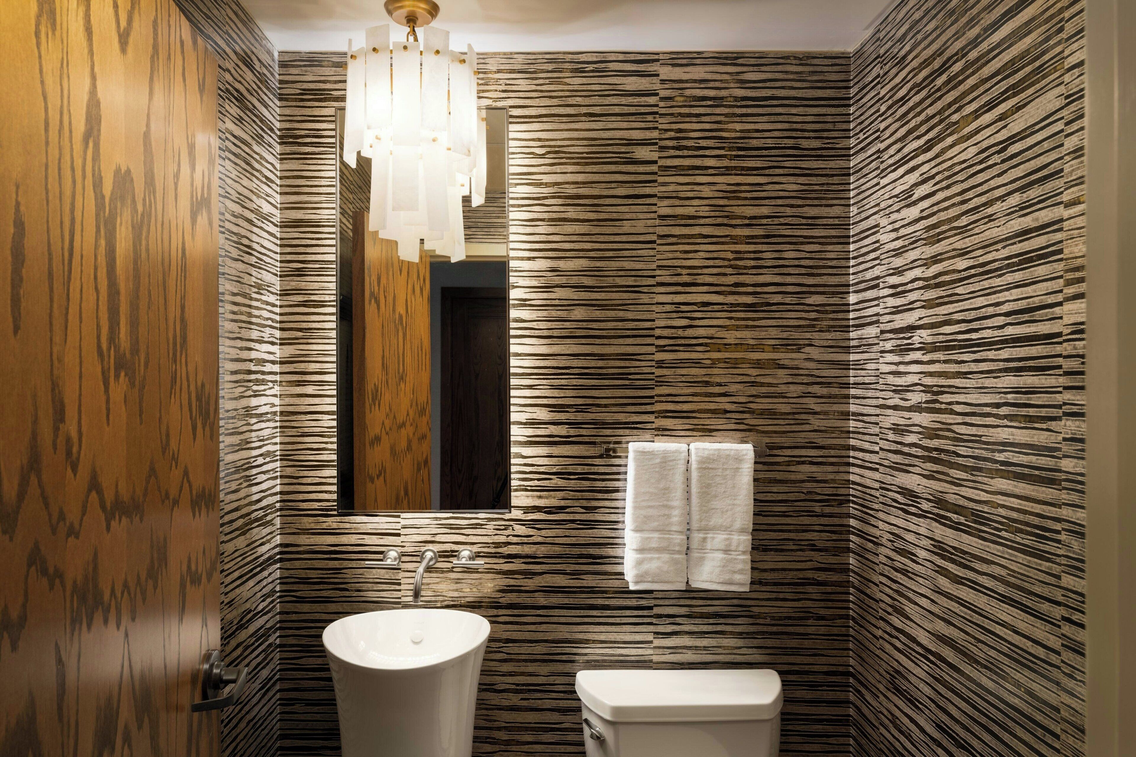 Ein Badezimmer mit Holztür, Waschbecken, Toilette, Spiegel und einer Kandelaberleuchte.