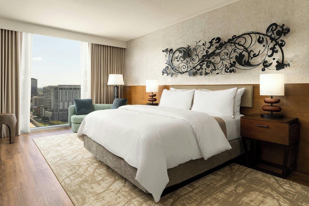 A hotel room with a large bed, two bedside tables with lamps, a decorative wall piece, and a view of the city through large windows.