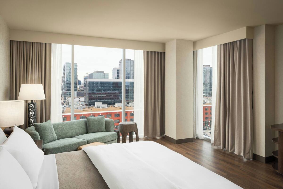 A hotel room with a bed, a green sofa, a small table, and a view of the cityscape through large windows.