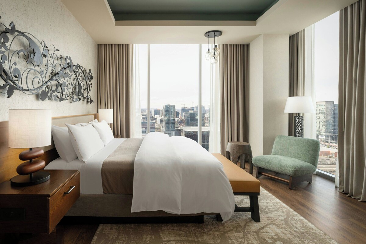 Habitación de hotel con una cama grande, un escritorio con lámpara, una silla y vista a la ciudad.