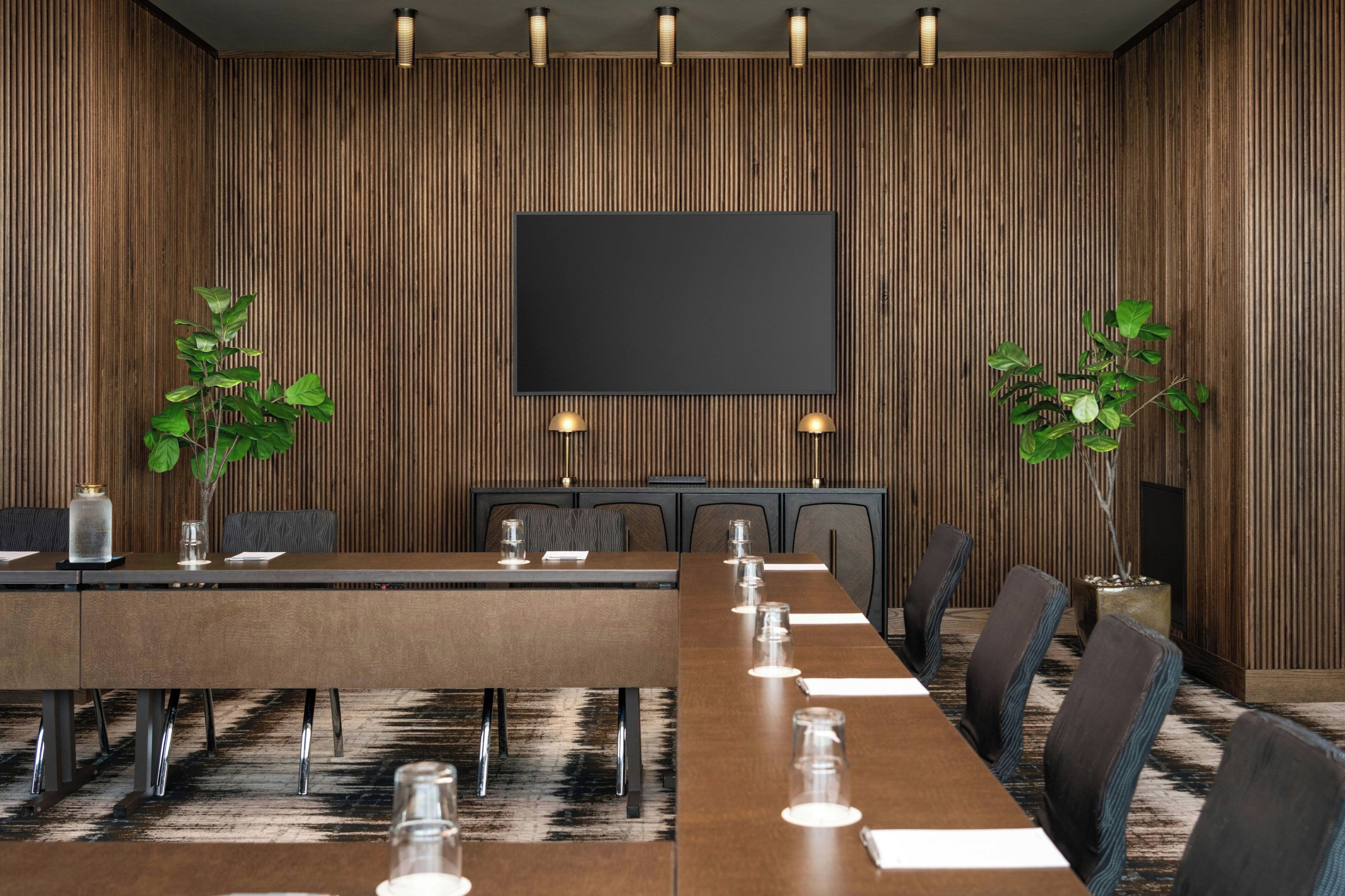 Un salón de conferencias con una larga mesa, sillas, una pantalla grande y dos plantas en macetas.