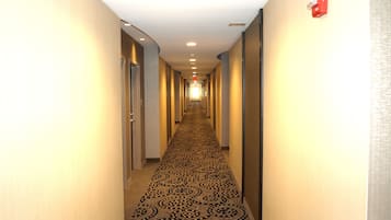 Pasillo de hotel con alfombra estampada, puertas a ambos lados y un detector de incendios rojo en la pared derecha.