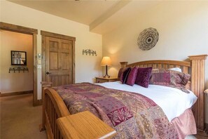 A bedroom with a wooden bed, a bedside table with a lamp, a decorative wall piece, and a door leading to another room. - 3106 Champagne Lodge, Trappeur's Crossing Apartment 2 SimplySteamboat (Steamboat Springs)