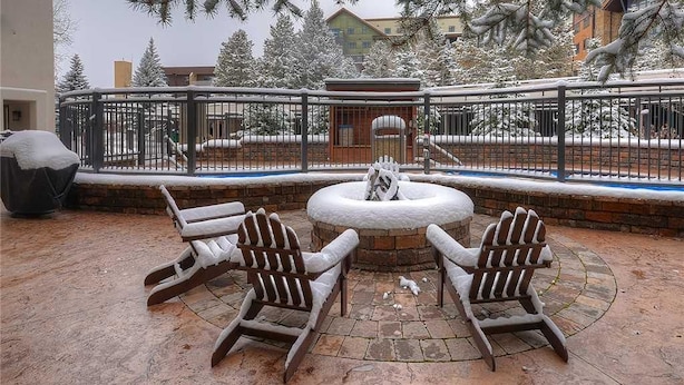 Un espace extérieur aménagé avec des sièges, recouvert de neige, comprenant des fauteuils Adirondack et un foyer, le tout entouré d’un mur de briques et d’une clôture métallique.