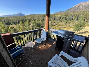 Una terraza con una parrilla, sillas y una mesa, con vista a una cadena montañosa boscosa.
