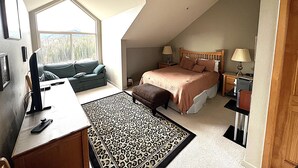 A bedroom with a bed, bedside tables, a desk, a chair, a sofa, and a window with a view of trees.