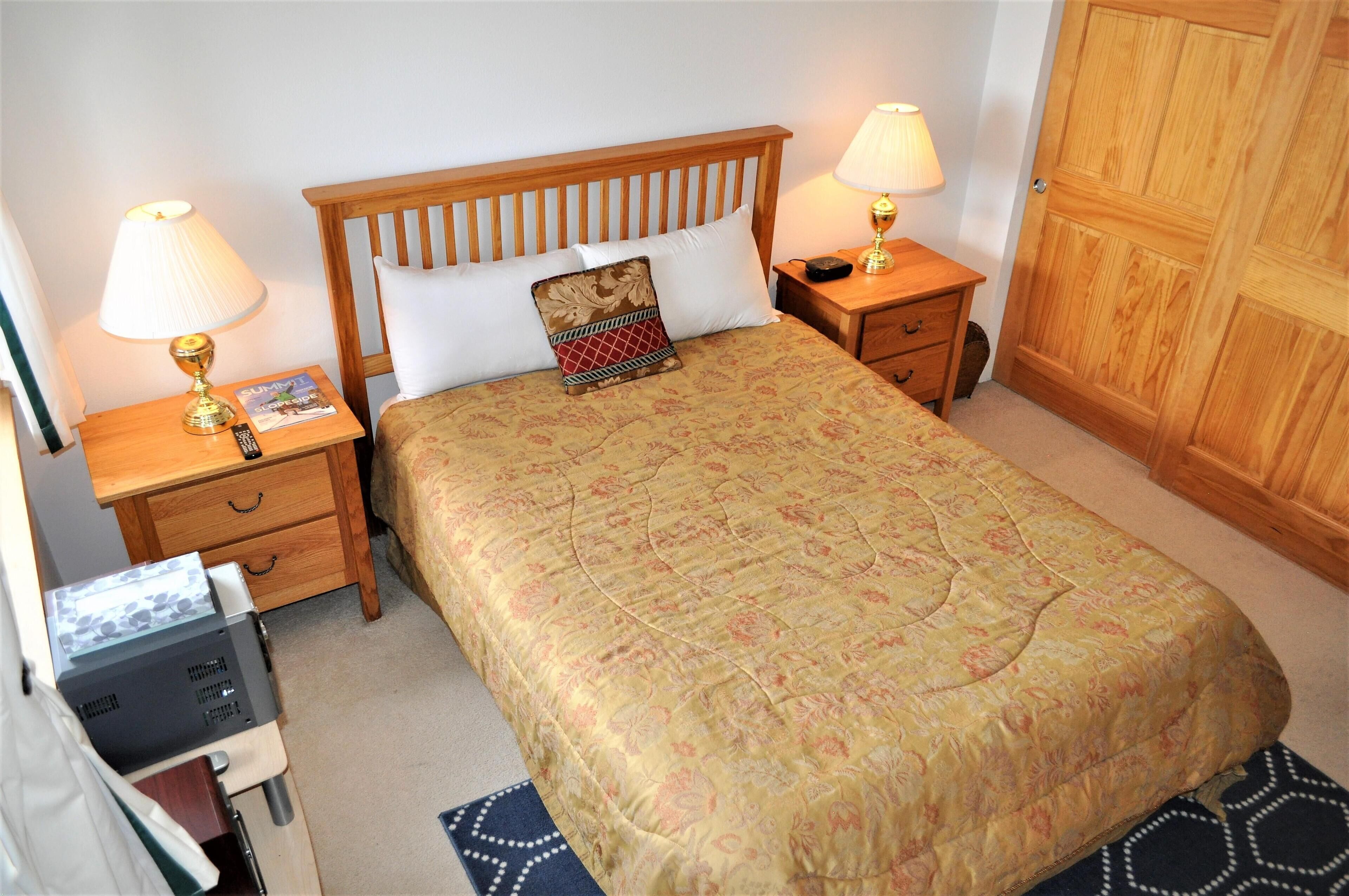 A neatly made bed with a floral bedspread, two bedside lamps, a wooden nightstand with a remote control, and a wooden wardrobe.