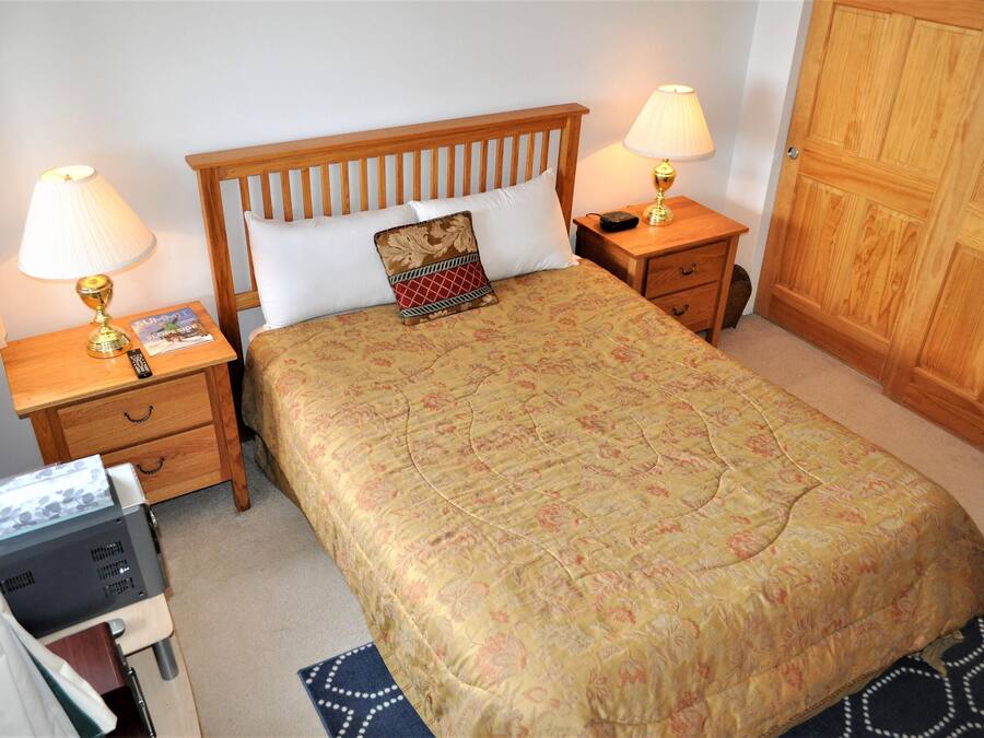 A neatly made bed with a floral bedspread, two bedside lamps, a wooden nightstand with a remote control, and a wooden wardrobe.