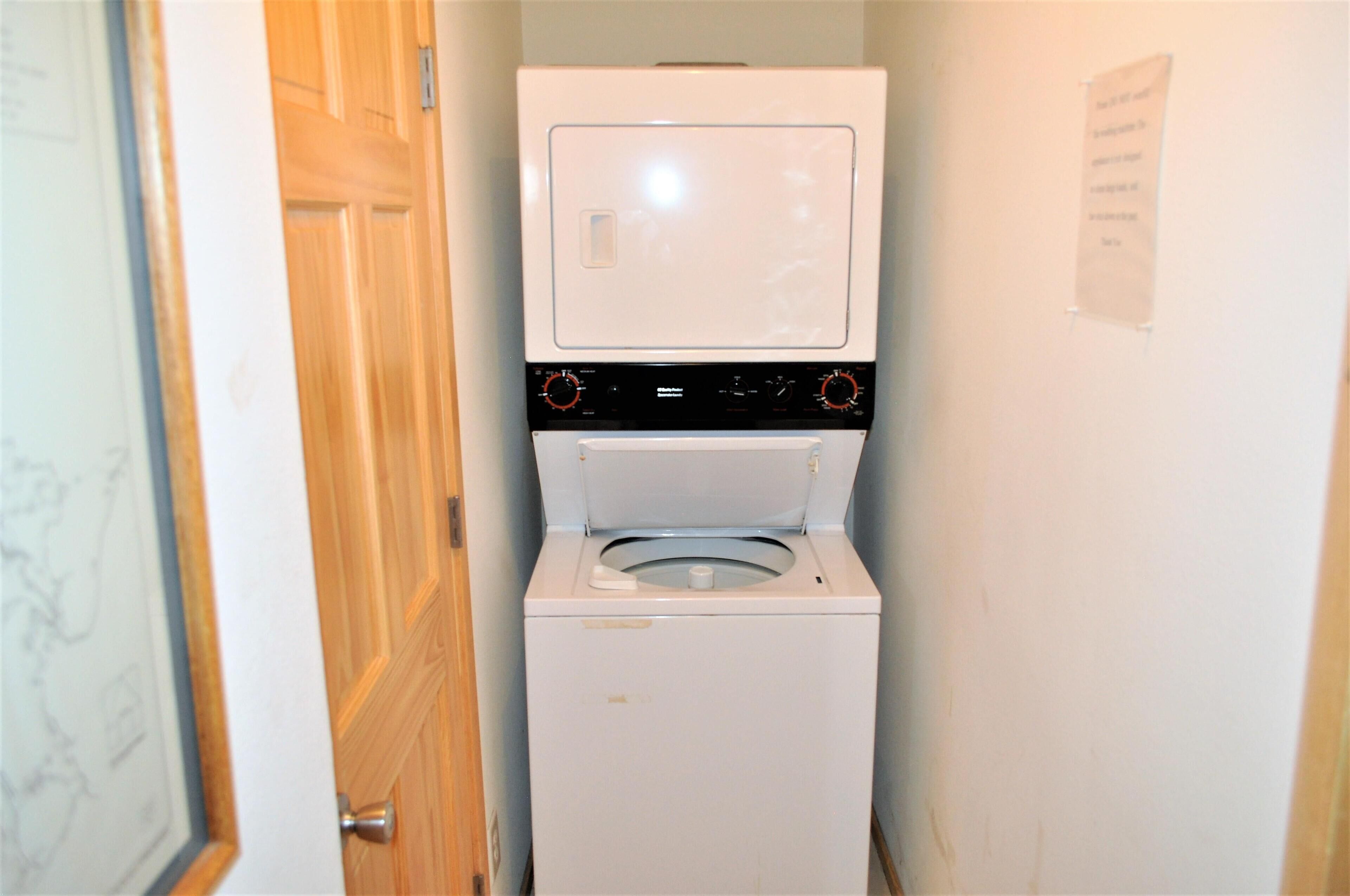 A compact laundry room with a front-loading washer and dryer, wooden door, and a wall-mounted sign.
