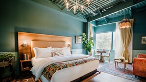 A modern bedroom with a large bed, a wooden headboard, a bedside table, a chair, a small table, and a potted plant. - The Frances Modern Inn (Austin)
