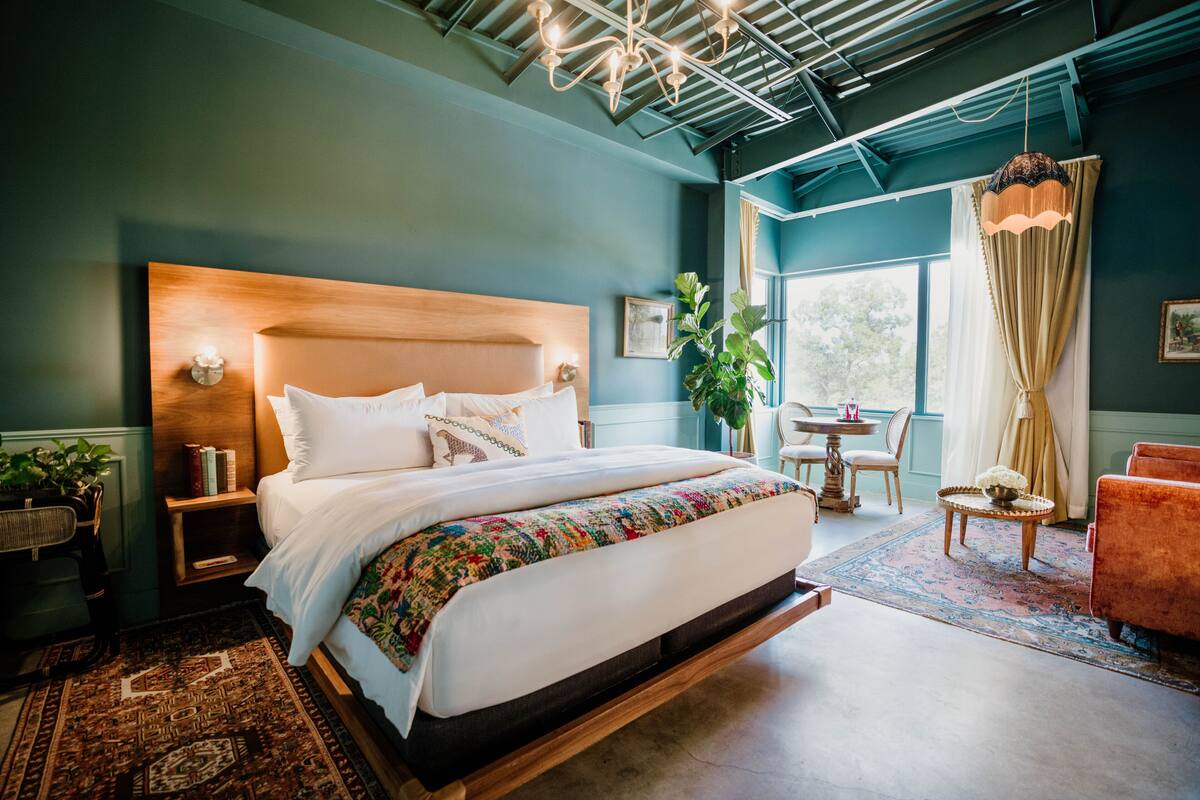 A modern bedroom with a large bed, a wooden headboard, a bedside table, a chair, a small table, and a potted plant.