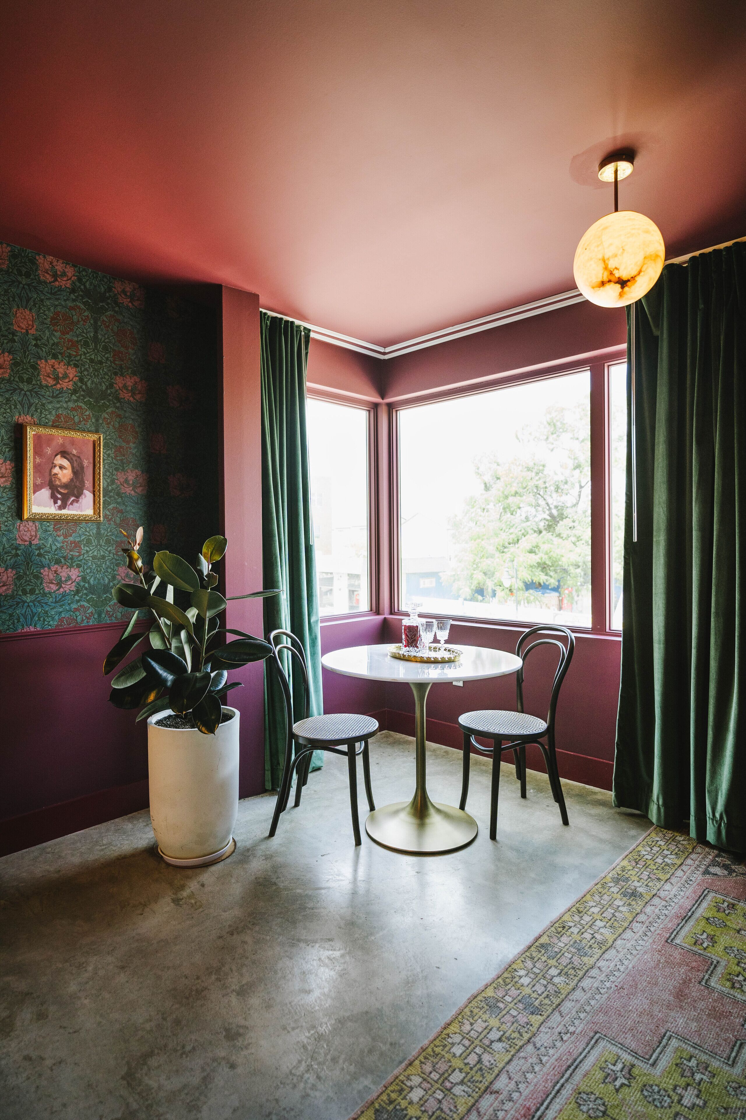 Una habitación con una mesa redonda, dos sillas, una planta en maceta, un cuadro en la pared y una ventana con cortinas.