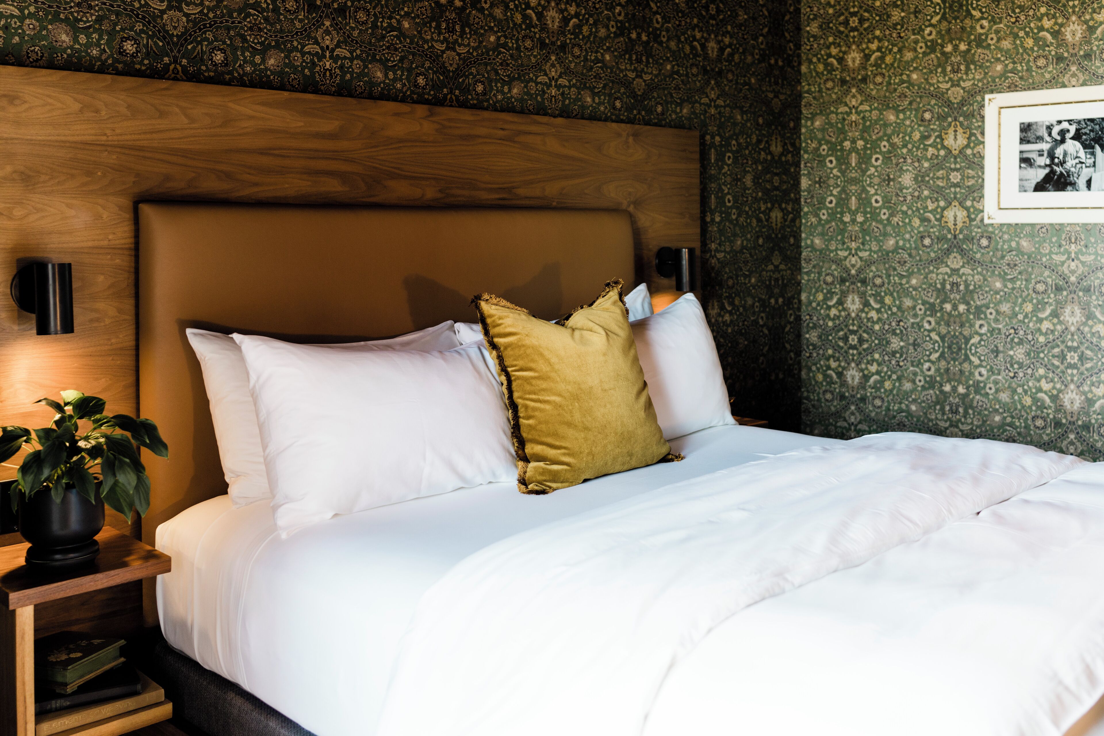 A neatly made bed with white and mustard pillows, a wooden headboard, and a nightstand with a plant.