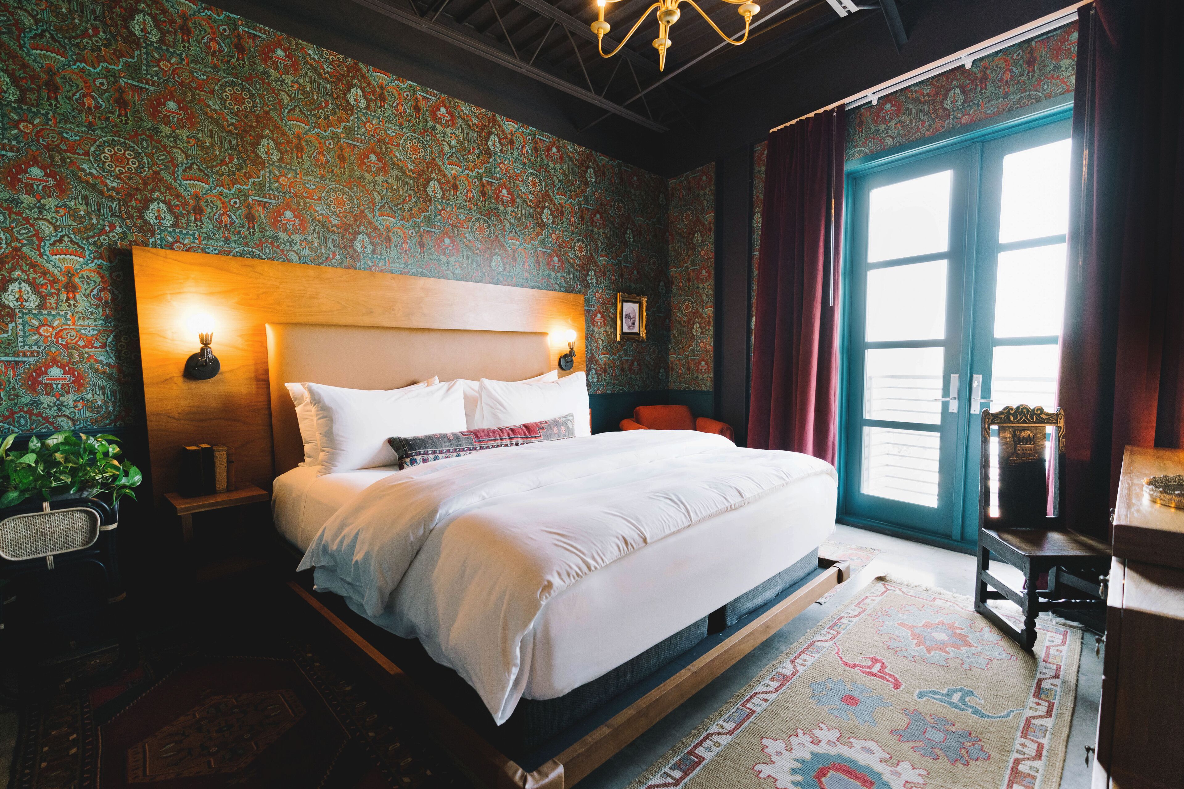 A bedroom with a large bed, patterned wallpaper, a wooden headboard, a nightstand, a chair, and a view of the outdoors through a window.