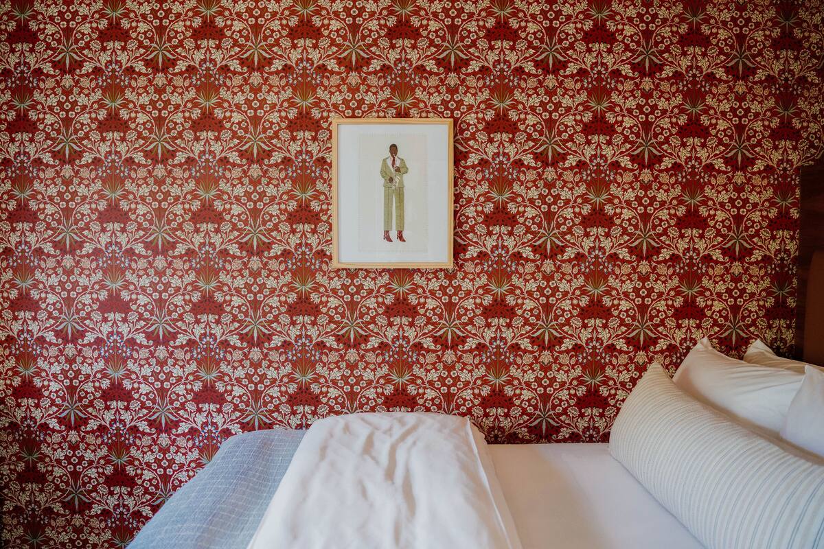 A hotel room with a patterned wallpaper, a framed picture on the wall, and a neatly made bed with pillows.