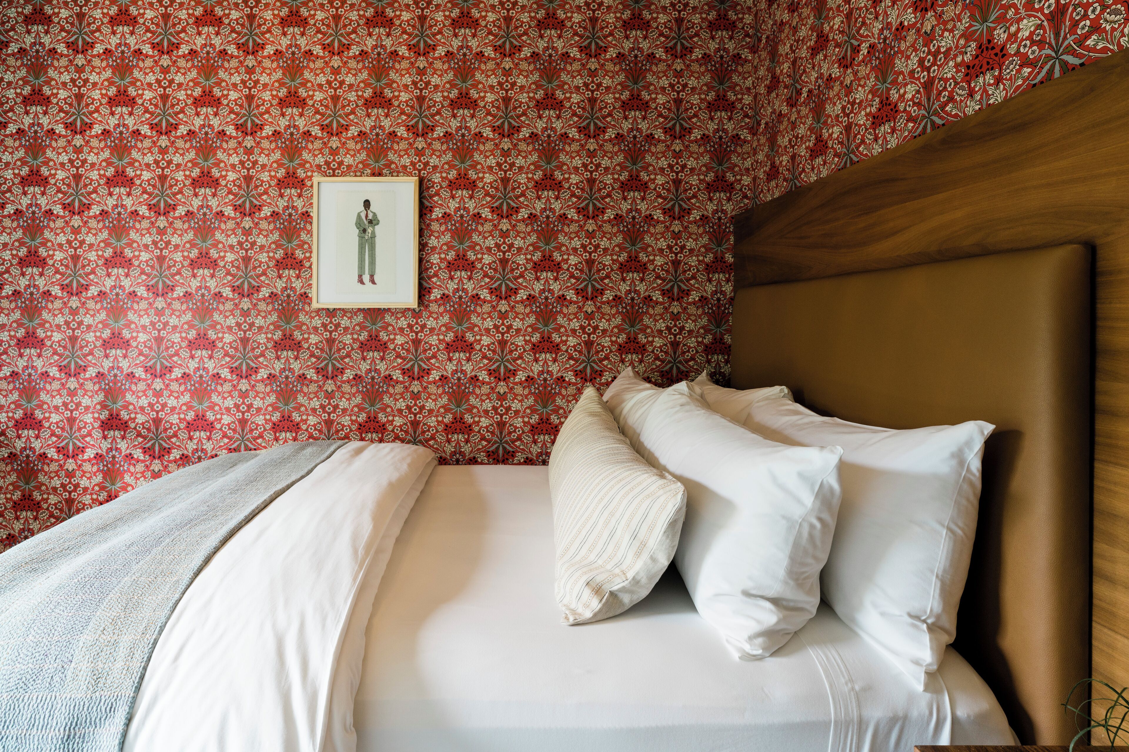 Una cama bien hecha con sábanas blancas y un plaid gris, un cabecero de madera y un papel tapiz floral rojo.