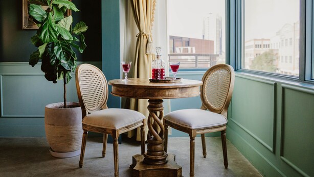 A round wooden table with two upholstered chairs, a potted plant, and two glasses of red drink on the table. - The Frances Modern Inn (Austin)