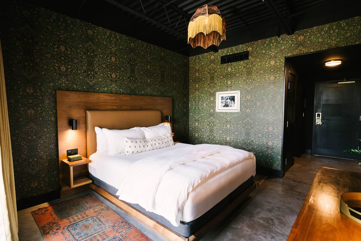 A neatly made bed with white linens, a wooden headboard, and a nightstand with a lamp. The room has patterned wallpaper and a large wooden door.