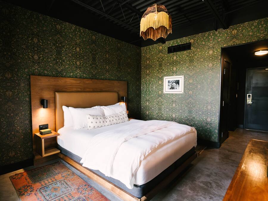 A neatly made bed with white linens, a wooden headboard, and a nightstand with a lamp. The room has patterned wallpaper and a large wooden door.