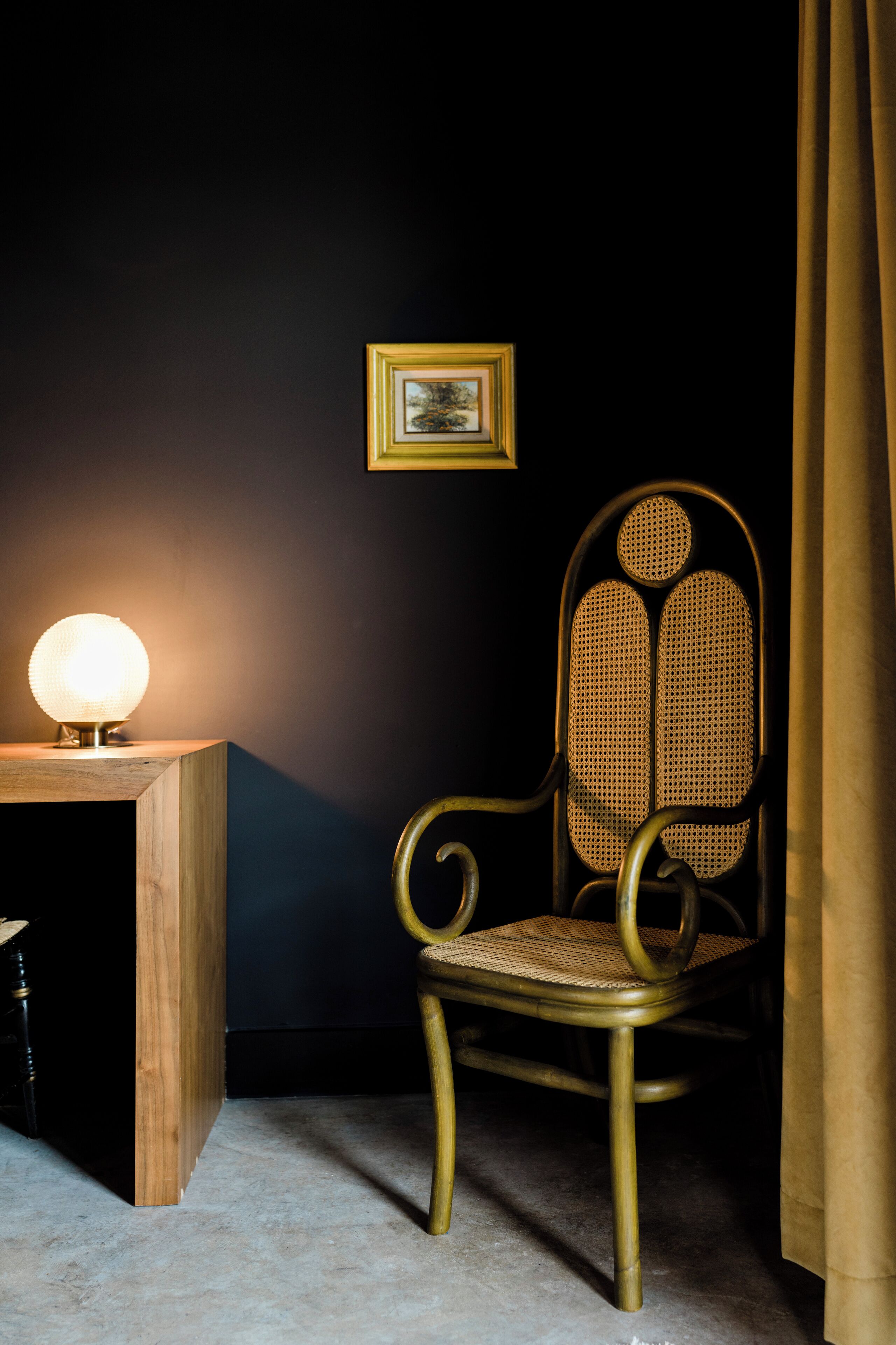 Una habitación con una mesita de madera, una lámpara, un sillón de mimbre, un cuadro en la pared y una cortina.