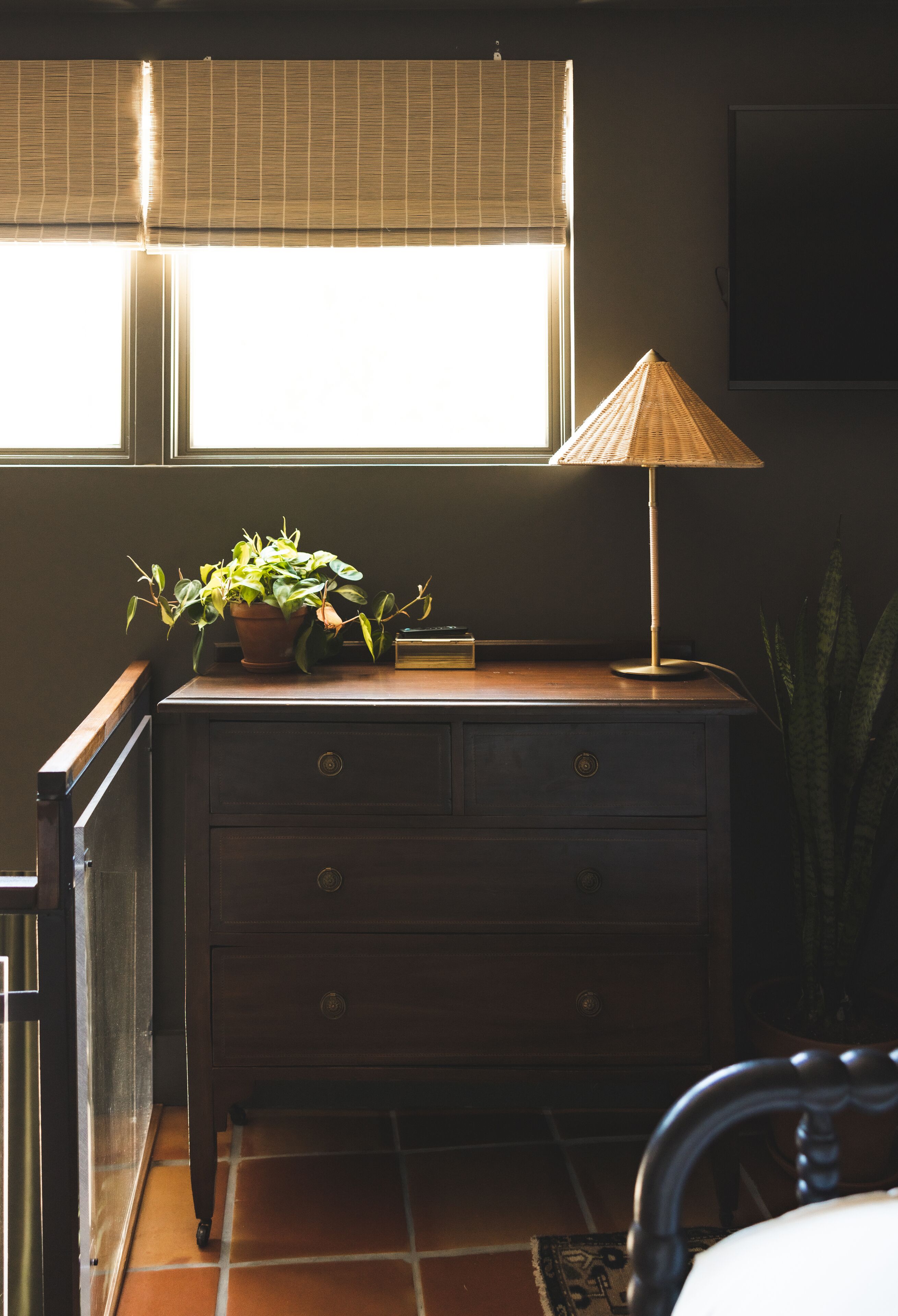 A room with a wooden dresser, a potted plant, a lamp, and a window with blinds.