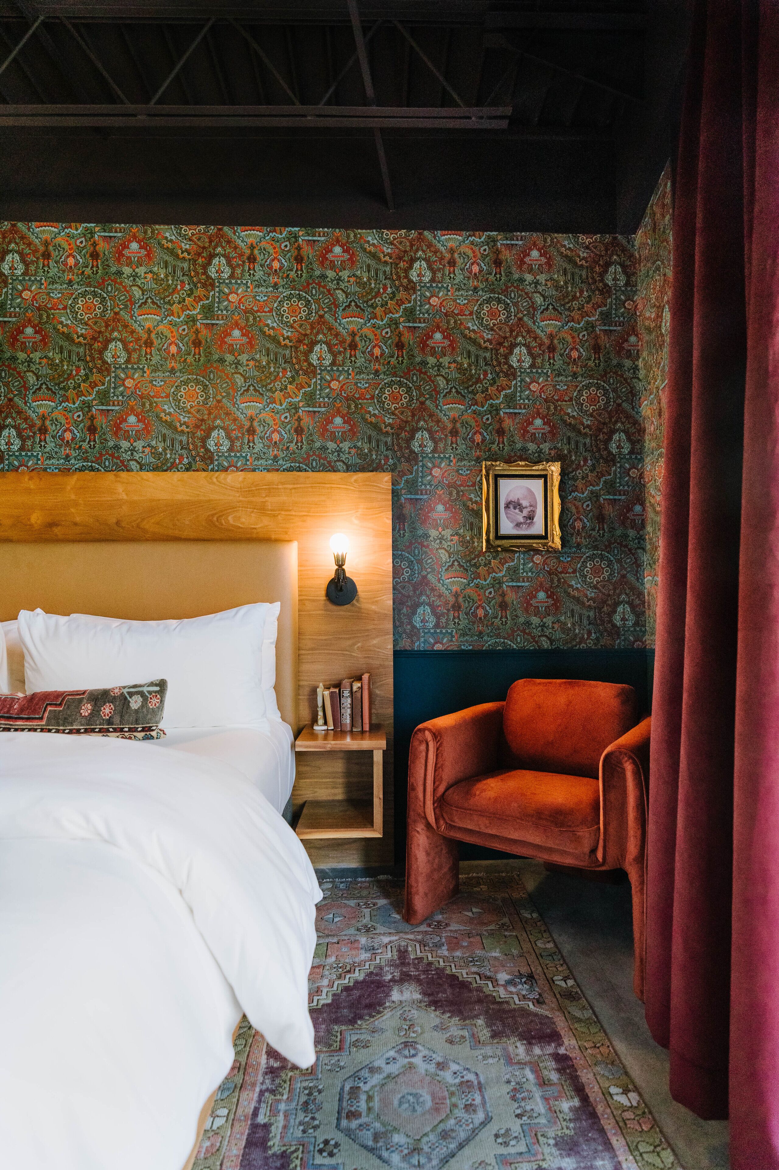Une pièce comprenant un lit, un fauteuil rouge, un tapis à motifs et un mur orné d’un tableau encadré.