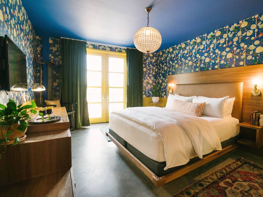 A bedroom with a large bed, a wooden headboard, a nightstand with a lamp, a potted plant, and a view of the outdoors through a window.