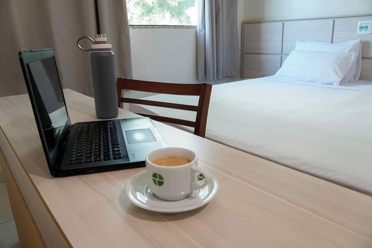A hotel room with a bed, a laptop, a water bottle, and a cup of coffee on a table.