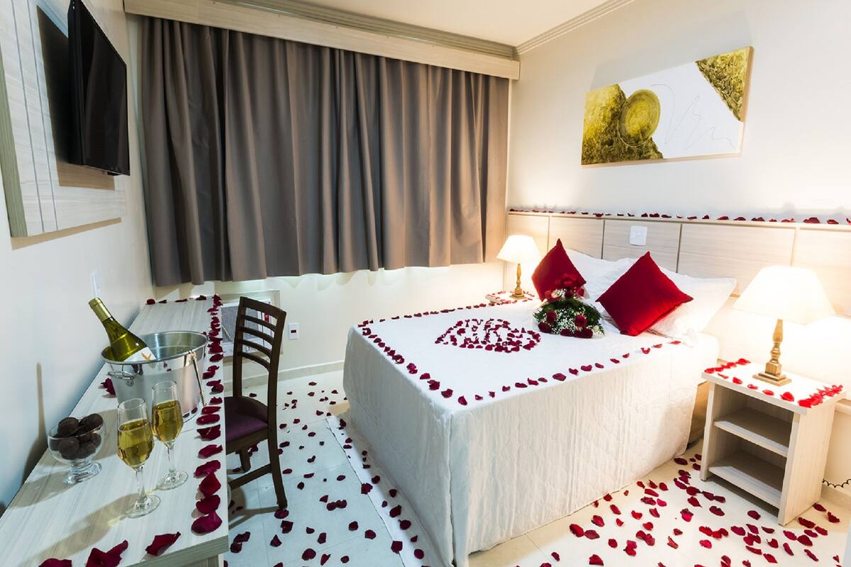 A hotel room with a bed, a bedside table, a chair, a small table with a bottle and glasses, and a wall with a painting.