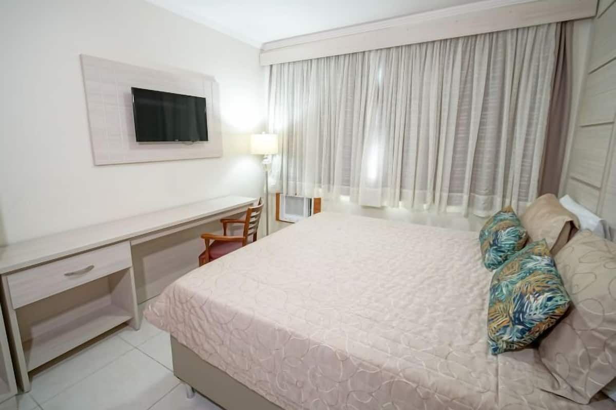A bedroom with a bed, a desk, a chair, a TV, and a window with curtains.