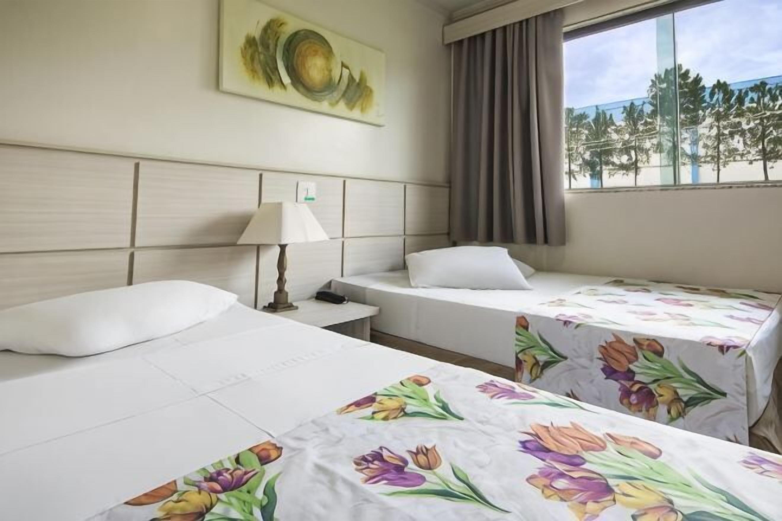 Een hotelkamer met twee bedden, een nachtkastje met een lamp, een schilderij aan de muur en een raam met uitzicht op bomen.