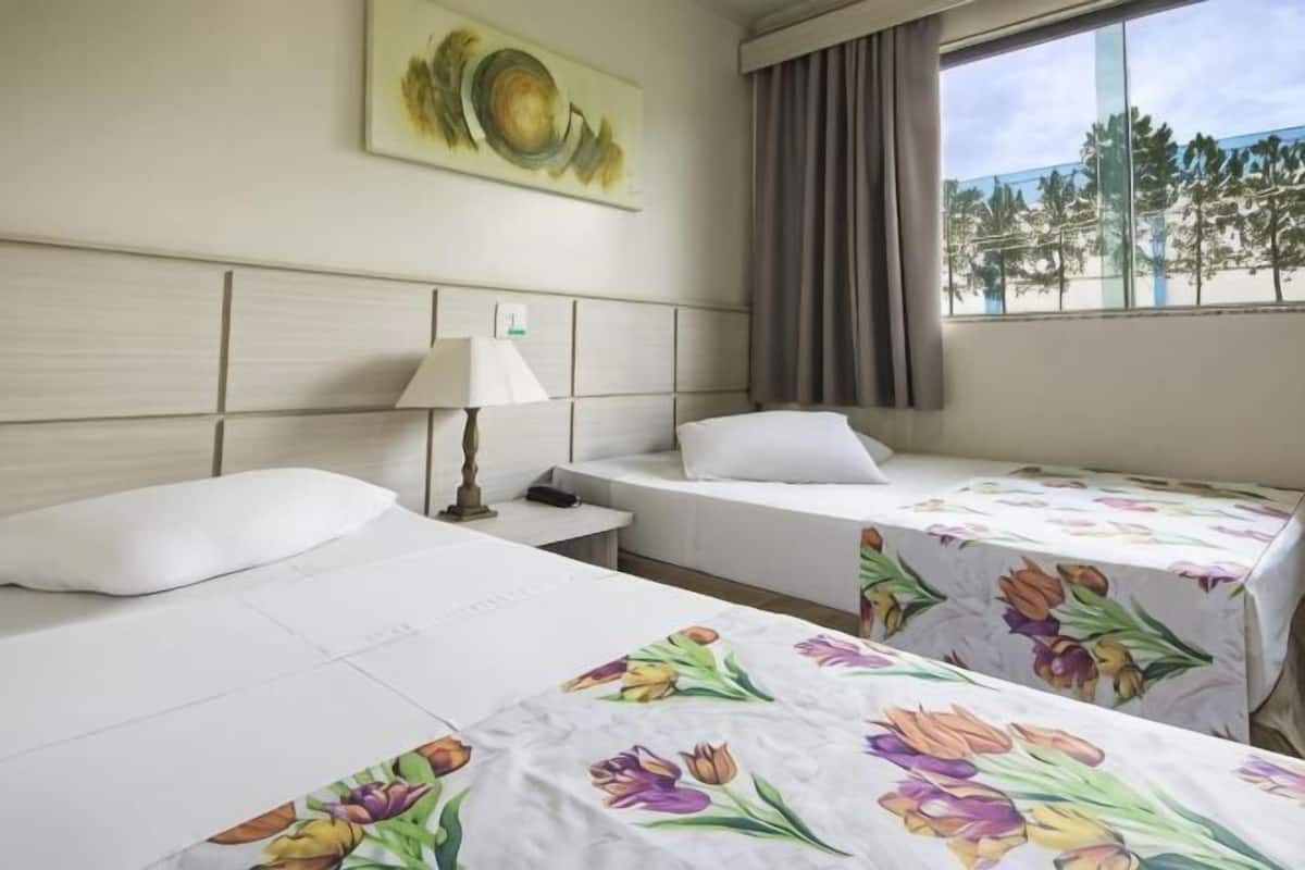 A hotel room with two beds, a nightstand with a lamp, a painting on the wall, and a window with a view of trees.