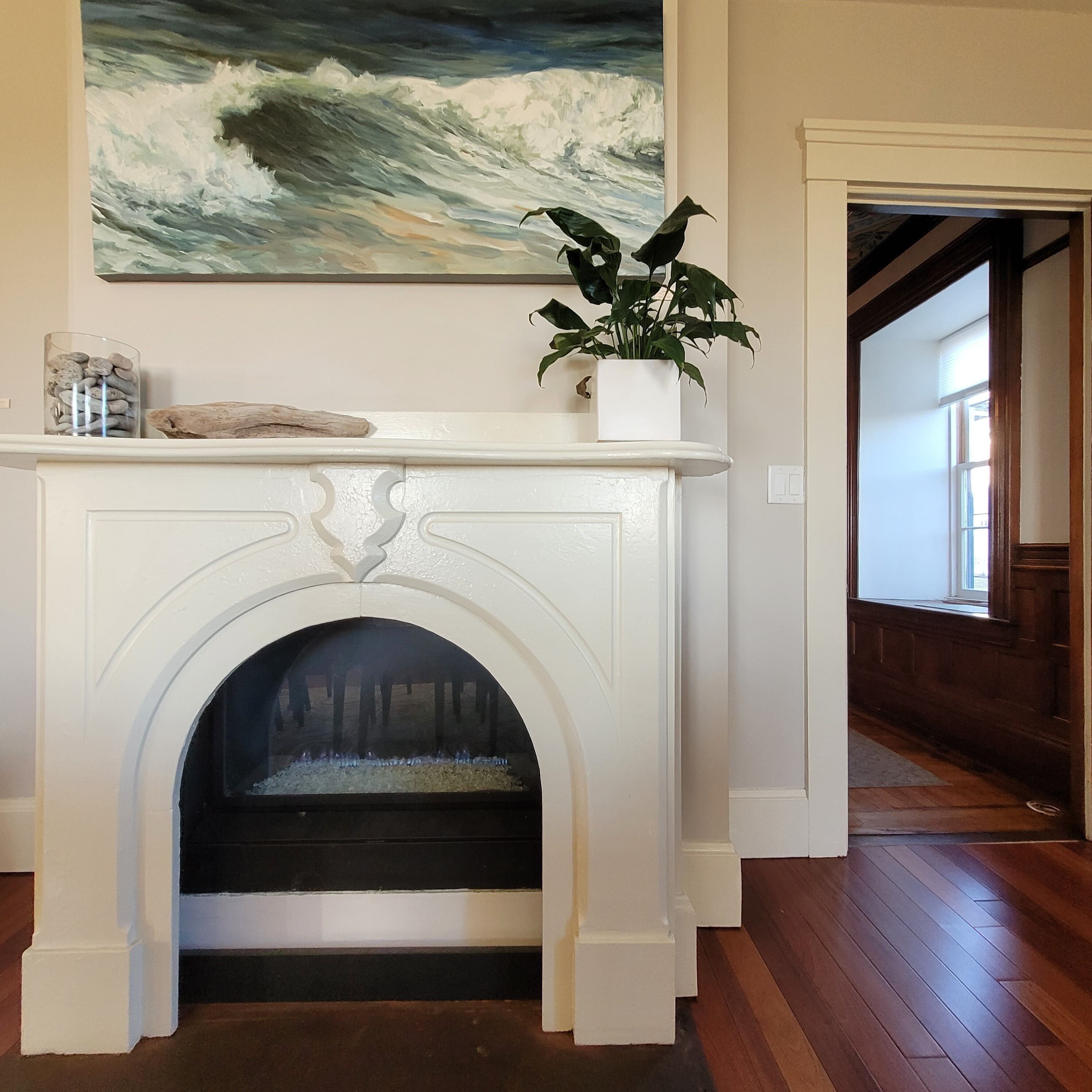 A white fireplace with a decorative arch, a potted plant on top, and a painting of waves above it.