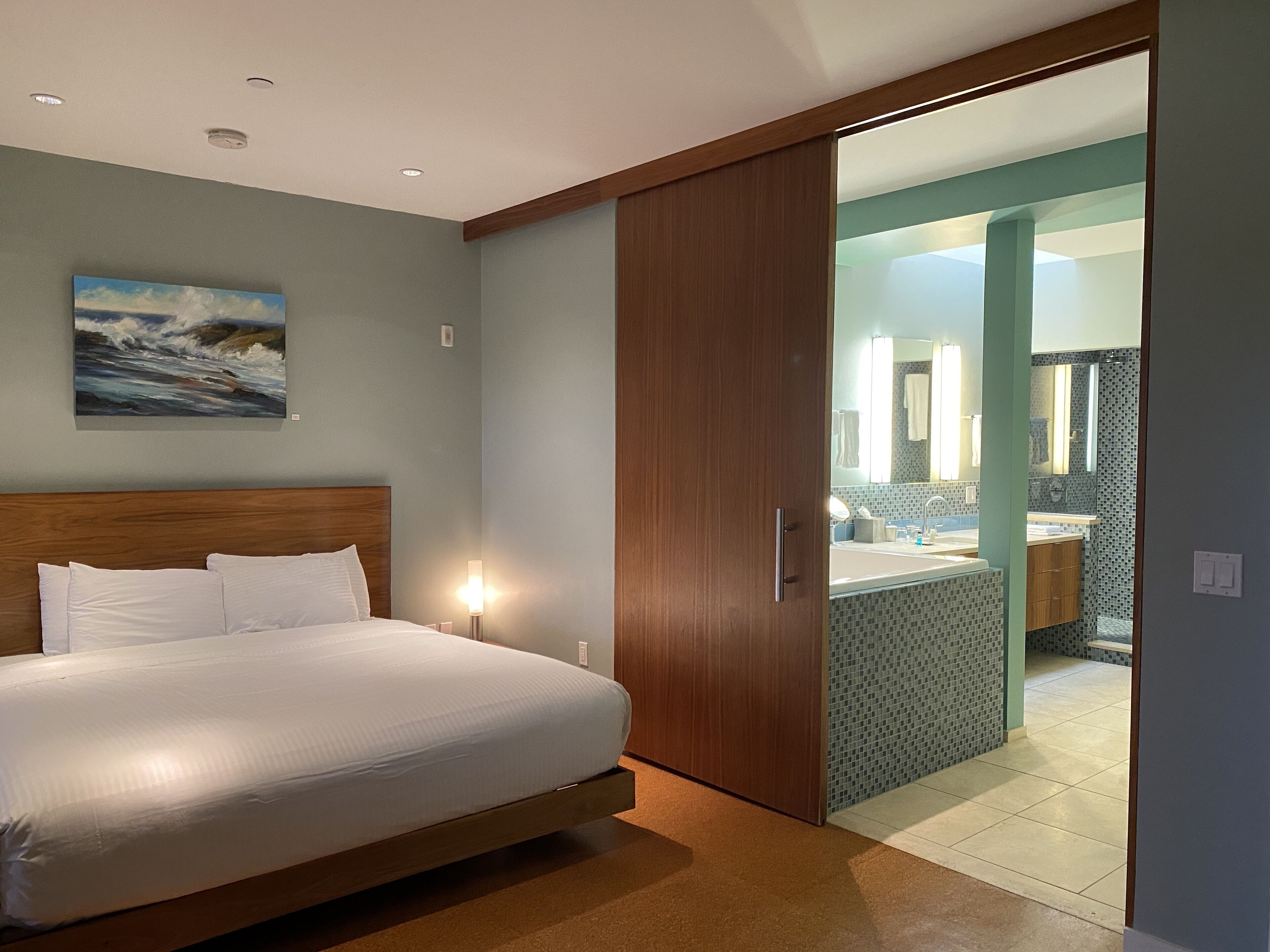 A bedroom with a bed, a painting, and a bathroom visible through an open door.