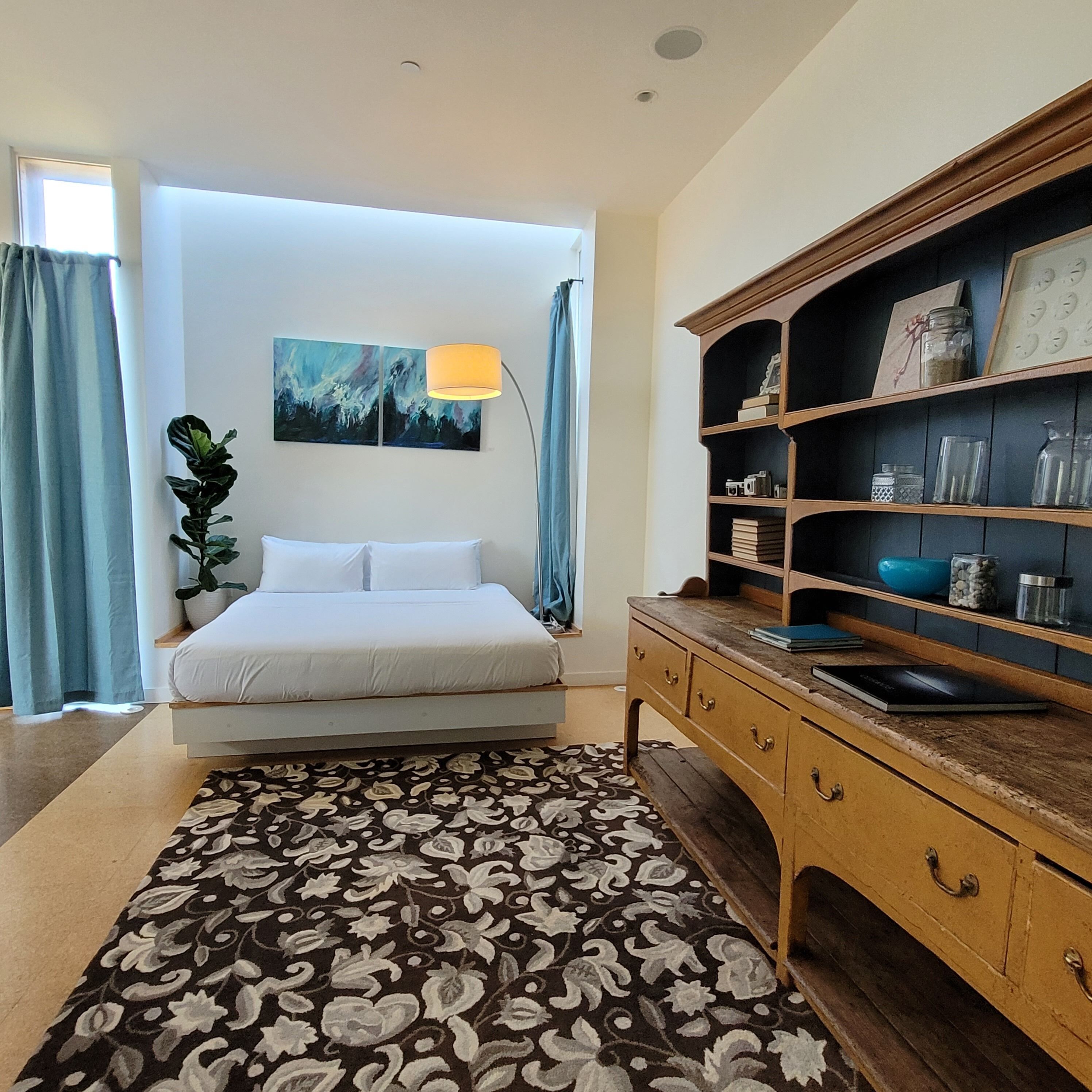 A bedroom with a bed, a bedside table, a lamp, a bookshelf, and a window with curtains.