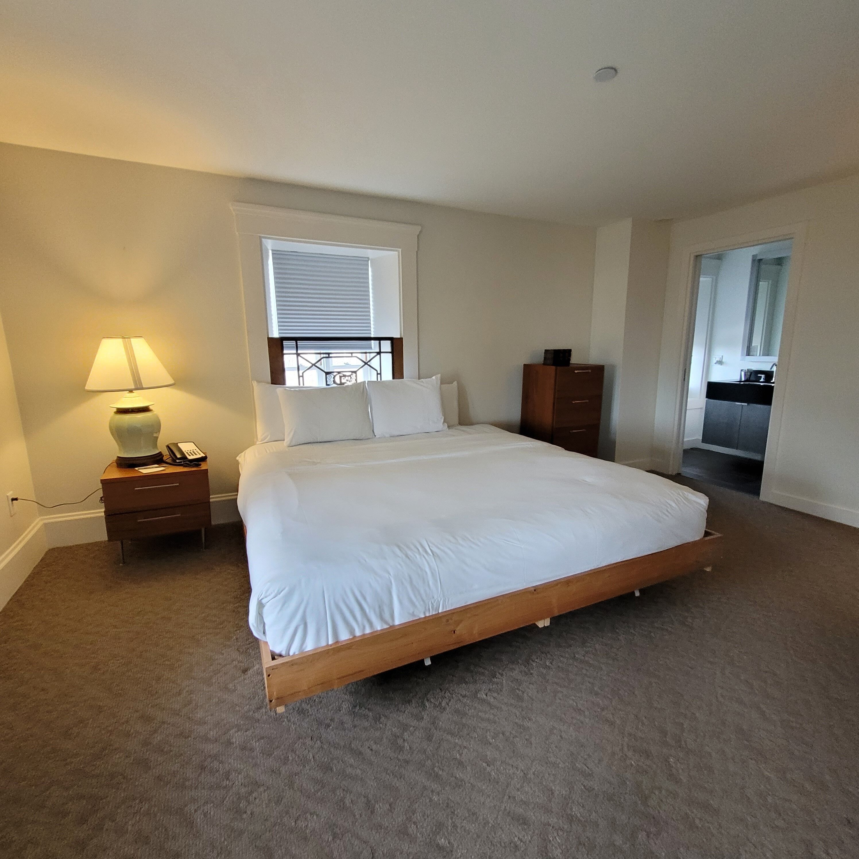 A bedroom with a single bed, a nightstand with a lamp, a wooden dresser, and a window with blinds.