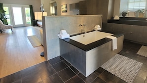 A modern bathroom with a large bathtub, a shower area, and a vanity. The room has a wooden floor and a large window providing natural light. - Stone House Inn (Little Compton)
