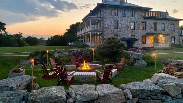 A large stone building with a wrap-around porch and a fire pit in the foreground. - Stone House Inn (Little Compton)